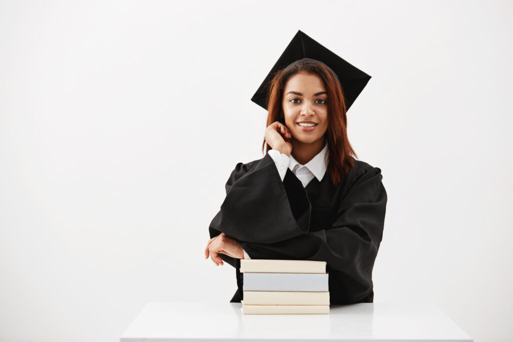 African female graduate in cap and mantle smiling sitting with books over white background.