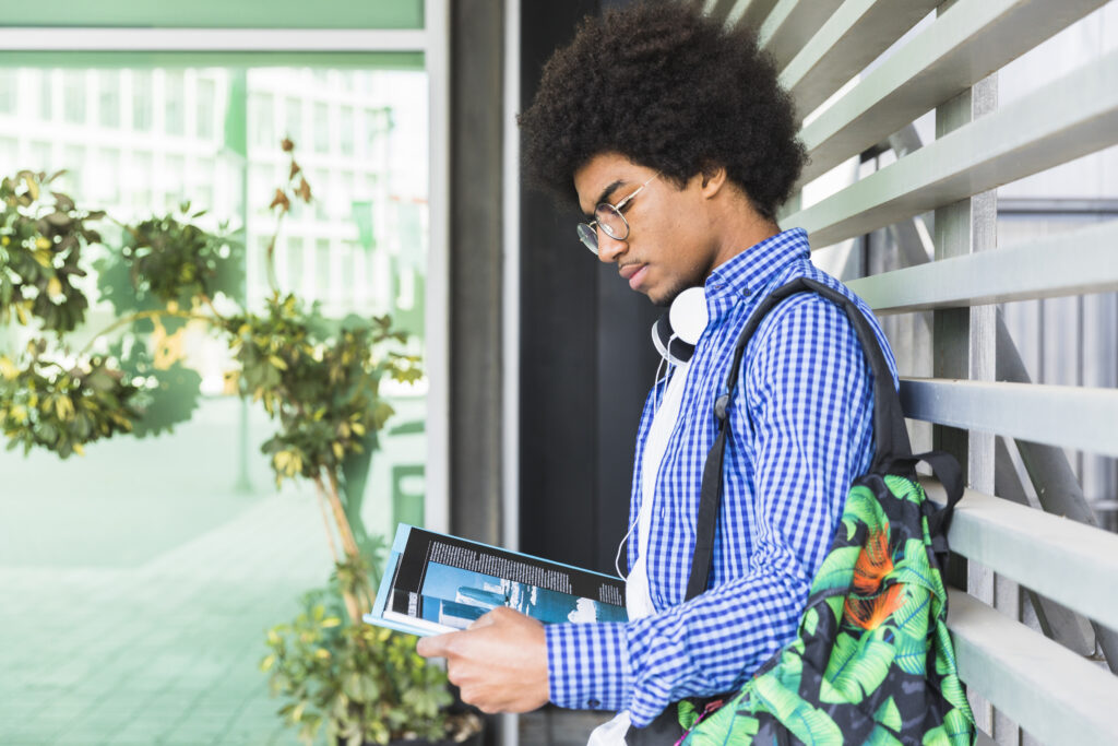 side-view-teenage-male-student-carrying-bag-his-shoulder-leaning-against-wall-reading-book